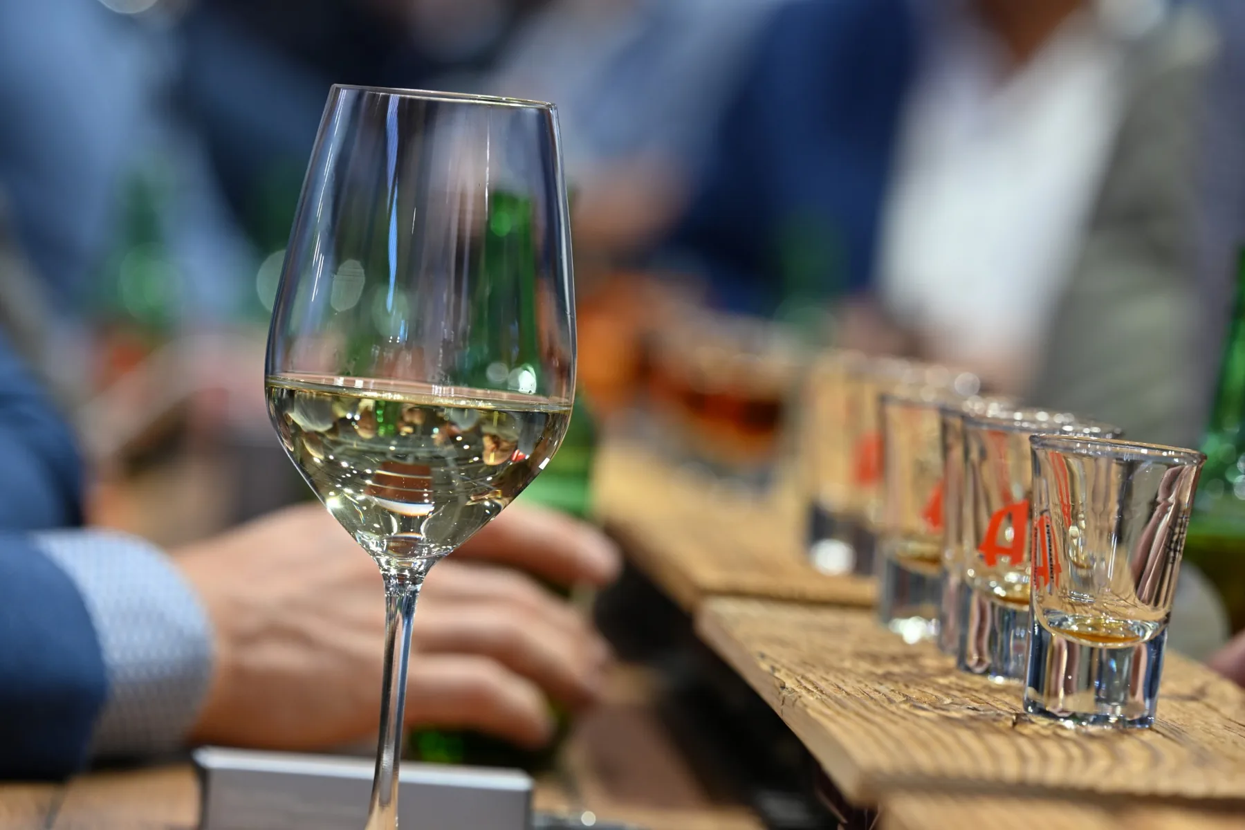 A glass of white wine and a row of shot glasses on the bar of the stand